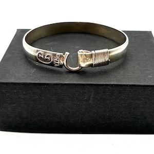 May include: A silver-toned bangle bracelet with a hook clasp. The bracelet has a smooth, rounded design with a decorative element near the clasp. The clasp is secured with a hook and loop closure. The bracelet is displayed on a black surface.