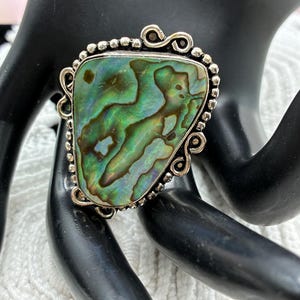 May include: A ring featuring a large, irregular, iridescent abalone shell inlay with swirling green, blue, and brown tones. The shell is set in a silver-toned metal frame decorated with small beaded details and decorative scrollwork accents.