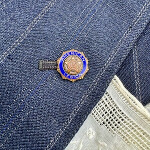 May include: A blue and gold American Legion pin is attached to a blue denim jacket. The pin has a blue border with the words "American Legion" and a gold centre with the letters "US". A white lace fabric is visible in the corner.