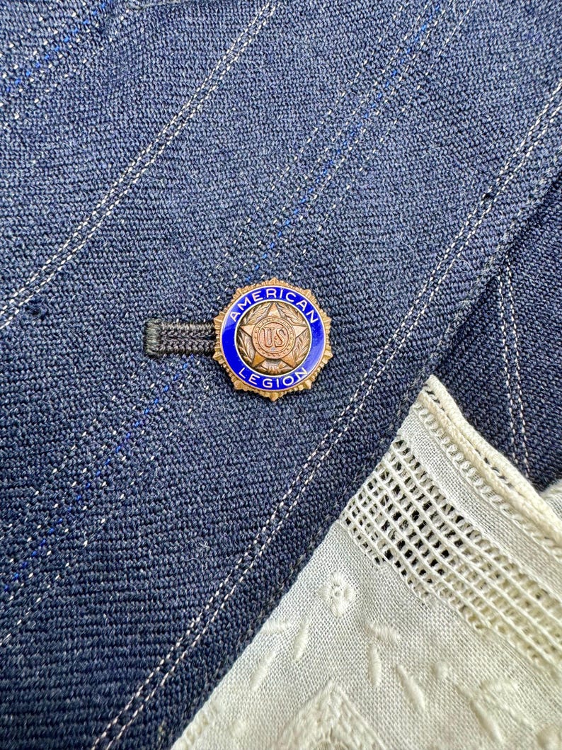May include: A blue and gold American Legion pin is attached to a blue denim jacket. The pin has a blue border with the words "American Legion" and a gold centre with the letters "US". A white lace fabric is visible in the corner.