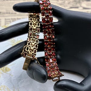 May include: A gold-toned bracelet featuring three parallel rows of oval-cut deep red garnet gemstones. The piece includes a secure metal clasp mechanism and a linked chain construction, showcasing a consistent pattern of faceted stones set in metal.