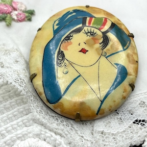 May include: An oval brooch featuring a hand-painted illustration of a person wearing a blue hat with red and green stripes. The figure has dark hair, long eyelashes, and a red diamond-shaped mouth. The brooch is set in a simple metal frame.