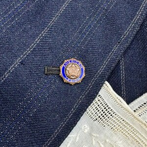 May include: A gold-coloured pin with a blue border and the words "AMERICAN LEGION US" is attached to a navy blue pinstripe blazer. A white lace handkerchief is visible in the lower right corner.