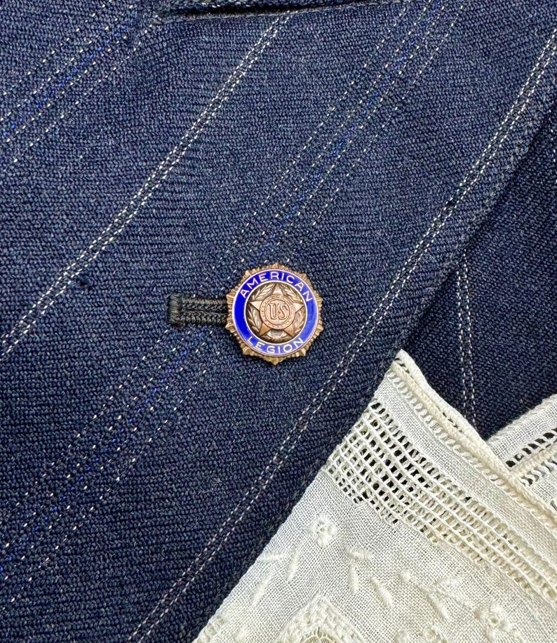 May include: A gold-coloured pin with a blue border and the words "AMERICAN LEGION US" is attached to a navy blue pinstripe blazer. A white lace handkerchief is visible in the lower right corner.