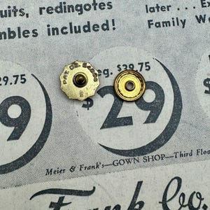 May include: Two vintage gold-coloured metal buttons with a circular design. One button has a starburst shape and the text "PAT. DE. 54295". The buttons are on a vintage newspaper background with text and the number "29".