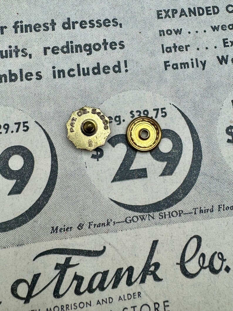 May include: Two vintage gold-coloured metal buttons with a circular design. One button has a starburst shape and the text "PAT. DE. 54295". The buttons are on a vintage newspaper background with text and the number "29".