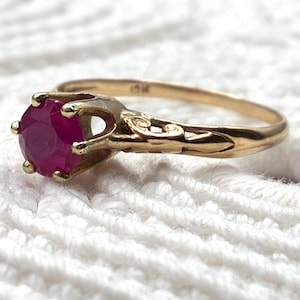 May include: A gold ring featuring a round, faceted, deep pink gemstone held in a six-prong setting. The band is crafted from yellow gold and displays intricate, decorative metalwork along the shoulders, with a visible 10K stamp on the inner band.