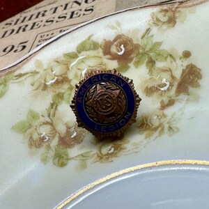 May include: A vintage American Legion pin with a blue and bronze design. The pin features the words "American Legion" around a central emblem. The pin is set against a floral patterned background.