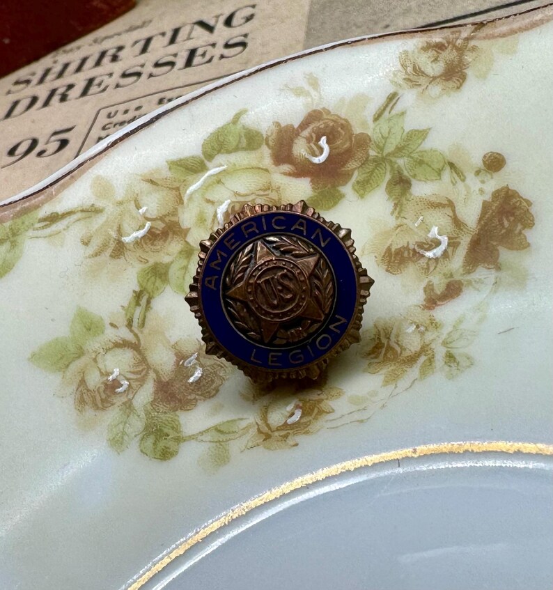 May include: A vintage American Legion pin with a blue and bronze design. The pin features the words "American Legion" around a central emblem. The pin is set against a floral patterned background.