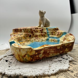 May include: A ceramic tray featuring a sculpted polar bear sitting on a rocky, textured landscape. The base has a mottled brown and tan finish with a recessed, glossy blue section representing water, creating a dimensional, landscape-like appearance.