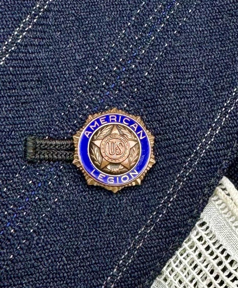 May include: Close-up view of a gold-colored American Legion lapel pin. The pin is circular with a blue enamel background.  The center features a gold star with the letters 'US' in a circular emblem.  The words 'AMERICAN LEGION' are inscribed around the edge. The pin is attached to a black backing.