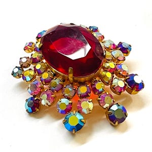 May include: Vintage brooch featuring a large, faceted, oval-cut ruby-red center stone. The brooch is surrounded by smaller, iridescent, multi-colored rhinestones in a gold-tone setting. A classic piece of jewelry.