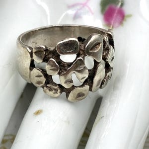 May include: A silver ring featuring an openwork design composed of multiple irregular, rounded, and polished metal segments. The band has a smooth, solid finish on the sides, while the center displays a textured, organic pattern with open spaces.