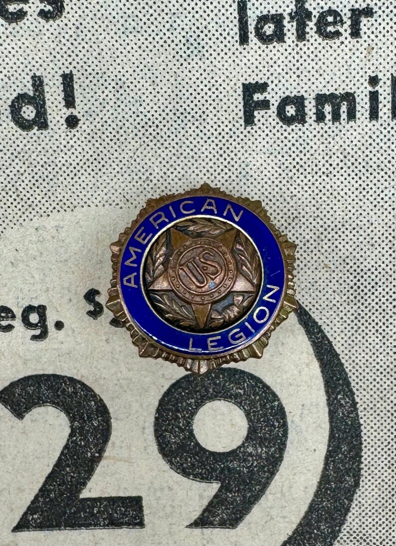 May include: A vintage American Legion badge with a bronze star and a blue enamel border. The badge features the words "AMERICAN LEGION" and "US" in the centre. The badge is circular and has a detailed design.