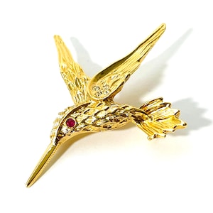 May include: A gold-colored hummingbird brooch with outstretched wings. The brooch features a ruby eye and small clear stones on the wings. The bird's feathers are detailed with textured patterns. The brooch is set against a white background.