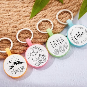 Op de afbeelding: Vier kleurrijke ronde sleutelhangers met zilveren ringen. Elke sleutelhanger heeft een ander ontwerp en een andere naam: Maggie, Stella, Layla en Chloe. De sleutelhangers hebben een witte achtergrond met zwarte illustraties en tekst.