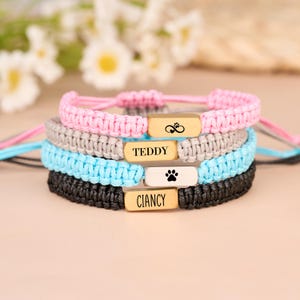 Könnte beinhalten: Vier Makramee-Armbänder in Rosa, Grau, Blau und Schwarz. Jedes Armband hat einen goldfarbenen rechteckigen Anhänger. Das rosa Armband hat einen Anhänger mit einem Pfotenabdruck und einem Herzsymbol. Auf dem grauen Armband steht "TEDDY". Das blaue Armband hat einen Pfotenabdruck. Das schwarze Armband sagt "CIANCY".