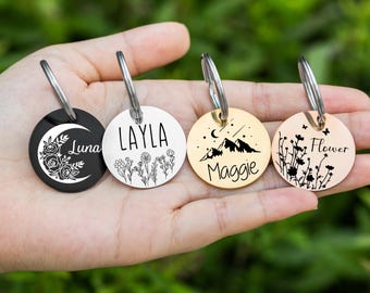 May include: Four round pet tags with silver key rings. One black tag features a crescent moon and floral design with the name "Luna". The other tags are gold-colored, with names "Layla", "Maggie", and "Flower" and various designs.