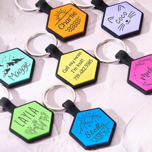 Può includere: Sette portachiavi esagonali colorati con diversi design e nomi. I portachiavi sono realizzati in un materiale resistente e presentano un anello nero per attaccare le chiavi. I nomi sui portachiavi includono Charlie, Coco, Maggie, Layla, Mocha, Stella e un numero di telefono.