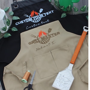 May include: Two aprons, one black and one tan, with embroidered text and logos. The black apron says "CUSTOM YOUR TEXT" and "Custom text". The tan apron says "GRILL MASTER" and "Mehmet K.". A spatula with an orange handle and the text "#1 MOM" is also visible.