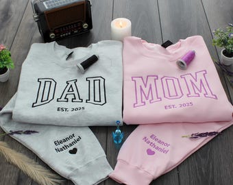 May include: A light gray sweatshirt with the word "DAD" in black and the text "EST. 2025". A pink sweatshirt with the word "MOM" in purple and the text "EST. 2025". Matching gray and pink sweatpants with names.