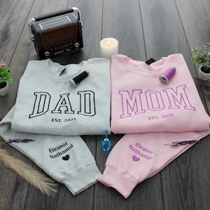 May include: Two gray and pink sweatshirts with the words "DAD EST. 2025" and "MOM EST. 2025" on them. The sweatshirts have the names "Eleanor Nathaniel" and a heart on the sleeves.