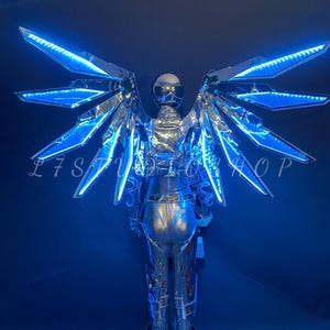 May include: A futuristic silver costume with illuminated blue wings. The costume includes a helmet and full body suit. The wings are made of reflective material and feature bright blue LED lights. The overall design is sleek and modern, perfect for a performance or cosplay.