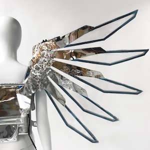May include: A futuristic, silver and clear wing-like costume piece. The wing has a complex, layered design with reflective surfaces and geometric shapes. The edges are outlined in a dark blue. The costume is attached to a mannequin.