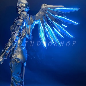 May include: A chrome-colored robotic costume with illuminated blue wings. The wings have a complex, layered design with embedded LED lights. The costume is highly reflective, and the image has the text "STUDIO SHOP".