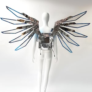 May include: A futuristic silver and blue wing costume displayed on a white mannequin. The wings have a layered design with reflective surfaces and blue trim. The costume is designed to be worn on the back.
