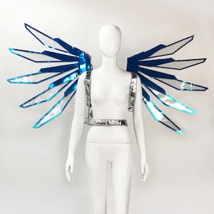 May include: A futuristic costume featuring a pair of blue and silver wings. The wings are attached to a silver harness worn by a mannequin. The wings have a geometric design with a translucent center and a bright blue outline. The mannequin is white and stands against a white background.