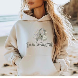 May include: A beige hoodie with a front pocket and a drawstring hood. The front of the hoodie features a design with a gray ribbon, floral elements, and the text "GLIO WARRIOR" in a script font. The hoodie is worn by a person.