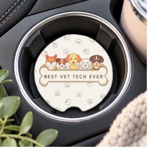 May include: A round car cup holder coaster with a cream background featuring a pattern of small gray paw prints. The center displays five cartoon dogs behind a large bone-shaped graphic with the black text BEST VET TECH EVER printed on it.