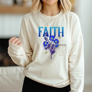 May include: Cream-colored long-sleeve shirt with the word "FAITH" in blue, featuring blue flowers, purple stems, and bees. The text "PSALM 18:2" and a Bible verse are also printed on the shirt.