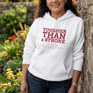 May include: A white hooded sweatshirt with a front pocket and the text "TOUGHER THAN a STROKE" in burgundy. The design includes a ribbon and stars. The person is wearing blue jeans and a silver watch.