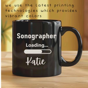 May include: Black coffee mug with white text that reads "Sonographer Loading... Katie". The mug has a white progress bar.