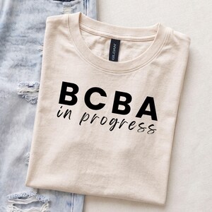 May include: A cream-colored short-sleeve t-shirt with a crew neckline and a black graphic print on the front. The text reads BCBA in a bold sans-serif font, followed by the words in progress in a smaller, cursive-style script font.