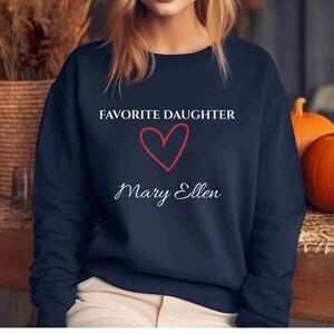 Puede incluir: Sudadera azul marino con el texto "FAVORITE DAUGHTER" sobre un coraz&oacute;n rojo y el nombre "Mary Ellen" en cursiva. La sudadera es de cuello redondo.