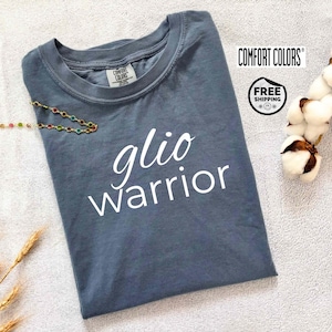 May include: A slate blue Comfort Colors t-shirt with the words "glio warrior" in white script. The shirt is folded and has a tag at the neckline. The image also includes a colorful beaded necklace and a sprig of cotton.