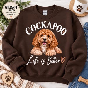 Peut inclure: Un sweat-shirt marron foncé à col rond avec une illustration d'un chien Cockapoo brun clair et poilu. Le texte au-dessus du chien indique COCKAPOO en blanc, et le texte en dessous indique Life is Better avec un petit cœur. Étiquette Gildan 18000.