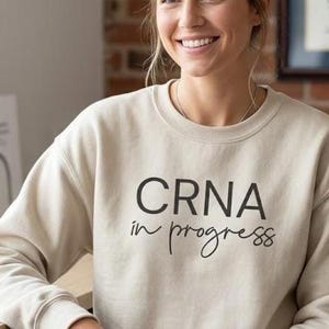 May include: A beige sweatshirt with the text "CRNA in progress" in black. The sweatshirt is worn by a person smiling. Books and a framed document are visible in the background.