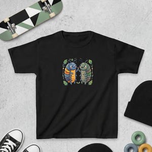 Puede incluir: Camiseta negra con una ilustración de dibujos animados de dos insectos tomados de la mano. Uno es azul y naranja, el otro verde y marrón, rodeados de hojas. La camiseta está hecha de material suave.
