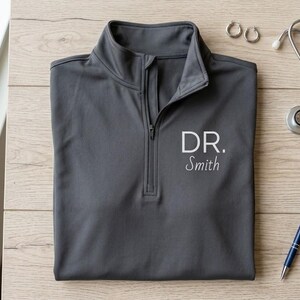 May include: A charcoal gray quarter-zip pullover shirt with the text "DR. Smith" embroidered in white. The shirt is laid flat on a wooden surface, accompanied by a stethoscope, pen, and earrings.