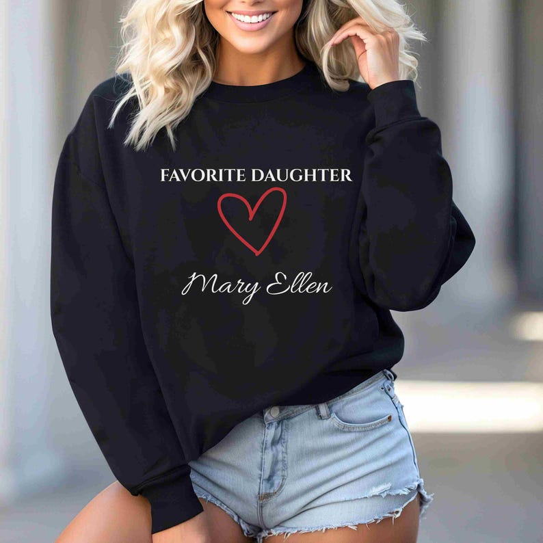 Puede incluir: Sudadera negra con el texto "FAVORITE DAUGHTER" sobre un coraz&oacute;n rojo y el nombre "Mary Ellen" en cursiva. La sudadera es de cuello redondo.