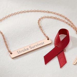May include: A rose gold necklace with the words "Stroke Survivor" engraved on a rectangular pendant. A red ribbon is next to the necklace. The necklace has a delicate chain and a small tag that says "Tees & Chic".