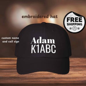 May include: Black baseball cap with the name "Adam" and call sign "K1ABC" embroidered in white. The text "embroidered hat" is above the cap. The words "custom name and call sign" are on the left side of the cap.