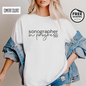May include: White Comfort Colors t-shirt with the text "sonographer in progress" in black script. The shirt is worn with a denim jacket and blue jean shorts. The shirt has a round neck and short sleeves.