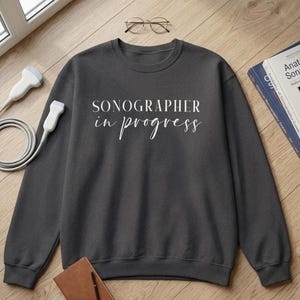 May include: A dark gray sweatshirt with the text "SONOGRAPHER in progress" in white script. The sweatshirt is on a wooden surface with a medical textbook, a brown notebook, and a pair of glasses. A medical ultrasound probe is also visible.