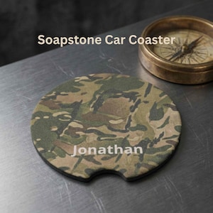 May include: A round soapstone car coaster featuring a camouflage pattern in shades of green, brown, and black. The name Jonathan is printed in white text across the centre. A small notch is cut out from the bottom edge of the coaster.