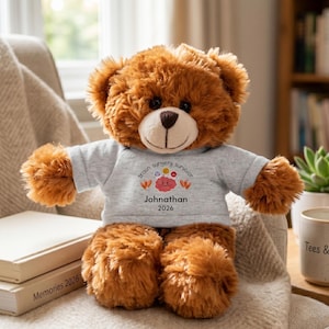 May include: A brown teddy bear wearing a gray t-shirt with the words "brain surgery survivor" and the name "Johnathan" and the year "2026". The bear is sitting on a beige blanket.